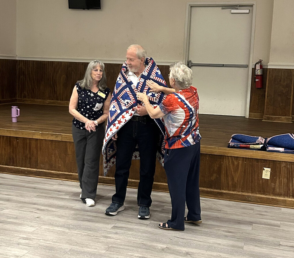 Women who created this quilt for Quilts of Valor wrapping a veteran recipient.